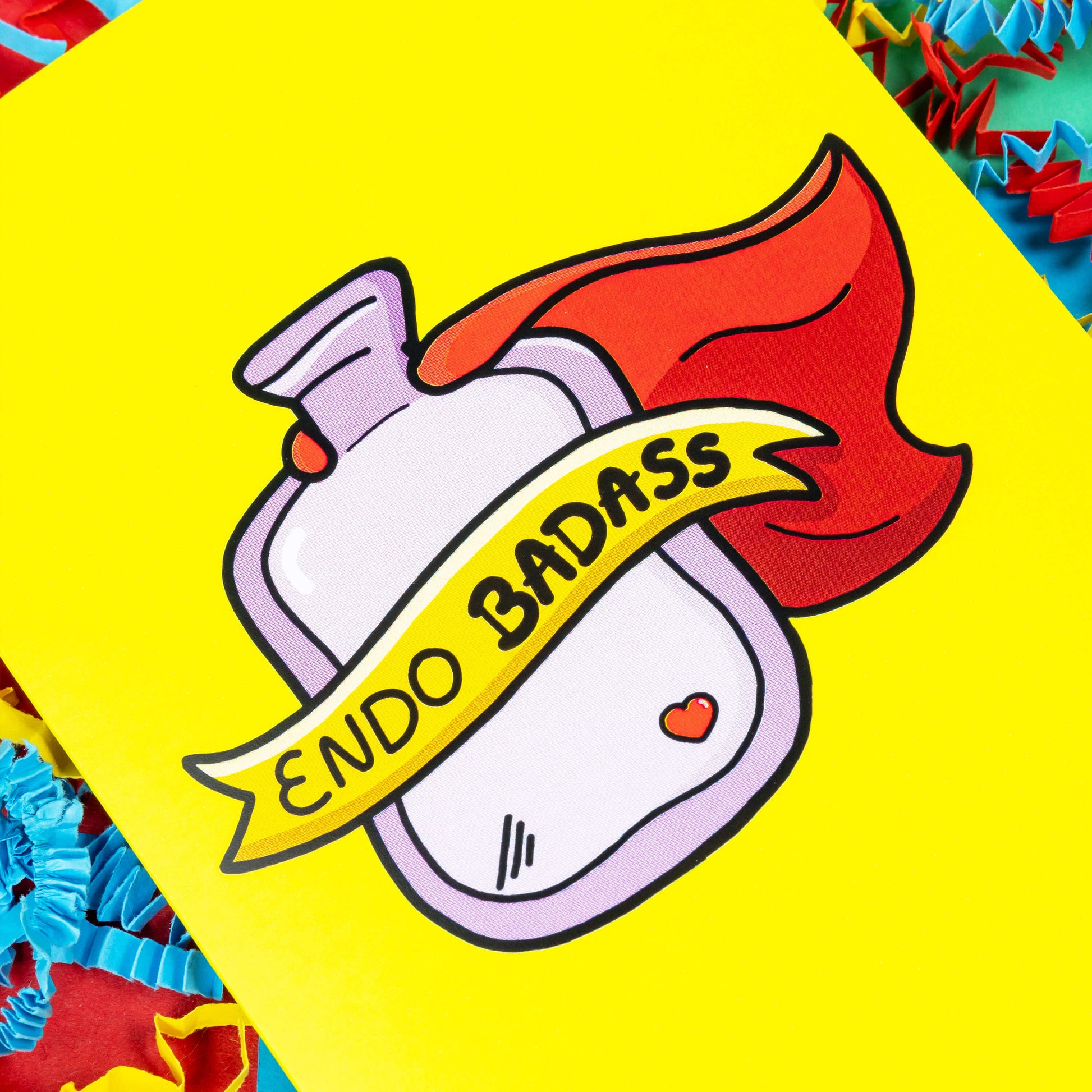 The Endo Badass Card - Endometriosis on a red, blue and green background with yellow, blue and red crinkle card confetti. The yellow based card features a pink hot waterbottle with a small red heart, a red cape and a yellow banner across reading 'endo badass'. Hand drawn design is raising awareness for Endometriosis and pelvic pain.