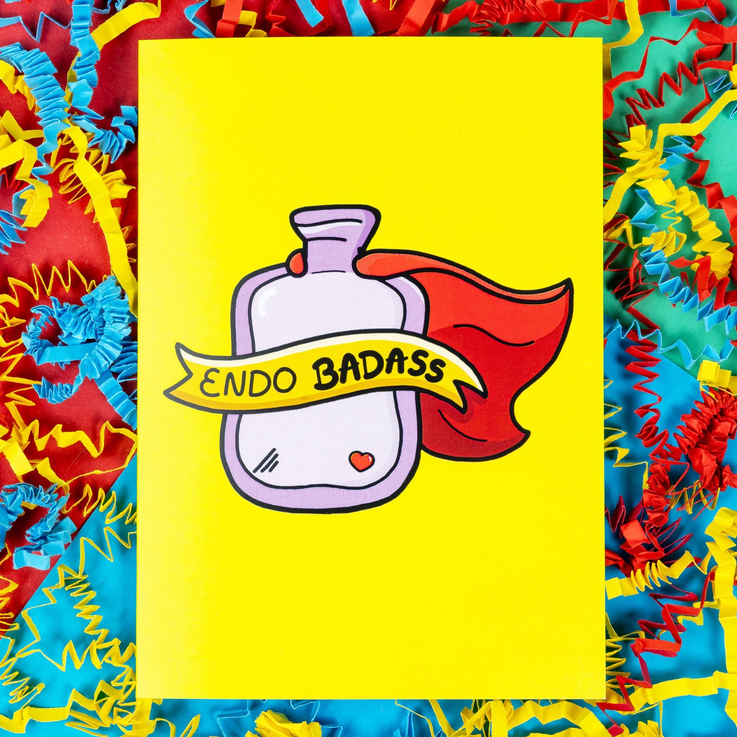 The Endo Badass Card - Endometriosis on a red, blue and green background with yellow, blue and red crinkle card confetti. The yellow based card features a pink hot waterbottle with a small red heart, a red cape and a yellow banner across reading 'endo badass'. Hand drawn design is raising awareness for Endometriosis and pelvic pain.