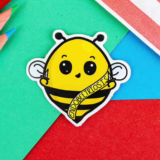 The Endombeetriosis Sticker - Endometriosis on a red, blue and green background with colouring pencils and red stripe candy bag. The yellow smiling bumble bee is holding daggers with a yellow sash ribbon reading 'endobeetriosis' in black. The hand drawn design is raising awareness for endometriosis.