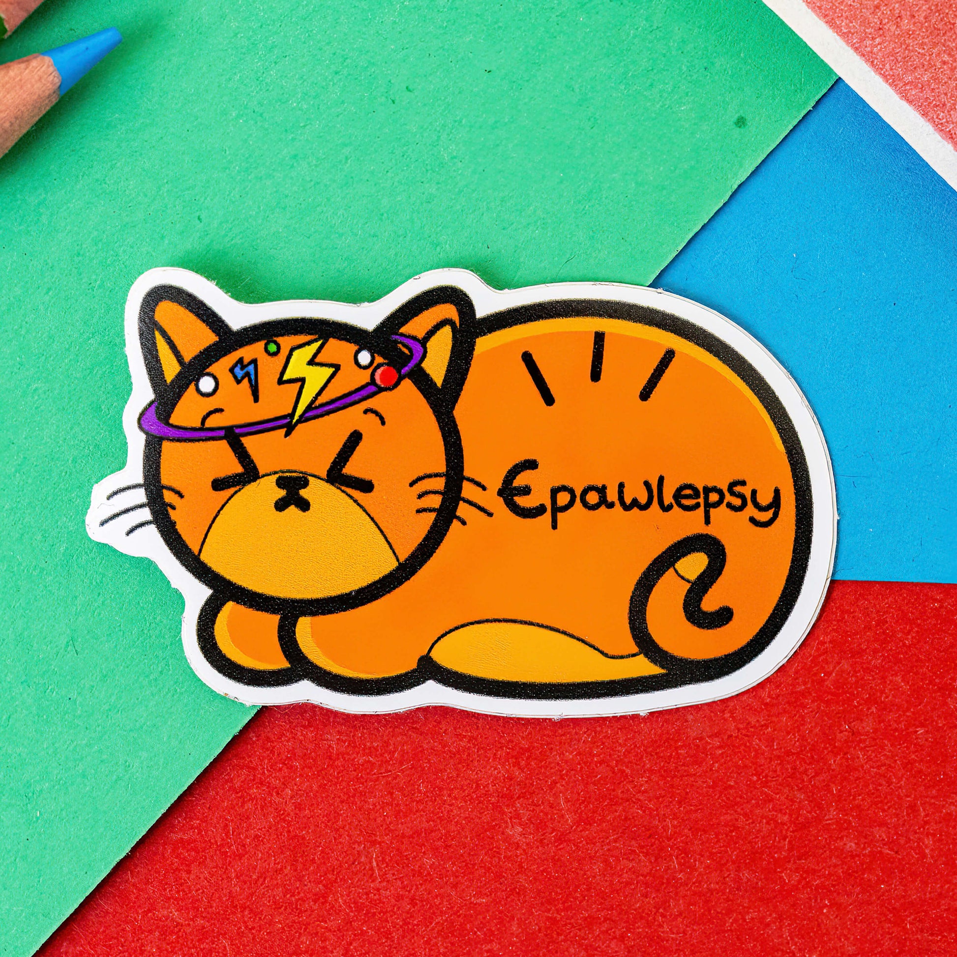 The Epawlepsy - Epilepsy Sticker on a red, green and blue background with colouring pencils. A ginger cat sticker with a dizzy spell drawn around its head it has the word 'Epawlepsy' written on it in black. The hand drawn design is raising awareness for epilepsy and seizures.