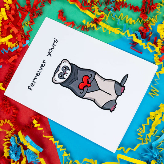 The Ferretver Yours Valentines Day Love Card on a red, blue and green background with red, yellow and blue crinkle card confetti. A white a6 greetings card with a drawing of a happy ferret holding a red heart saying ferreter yours on the front.