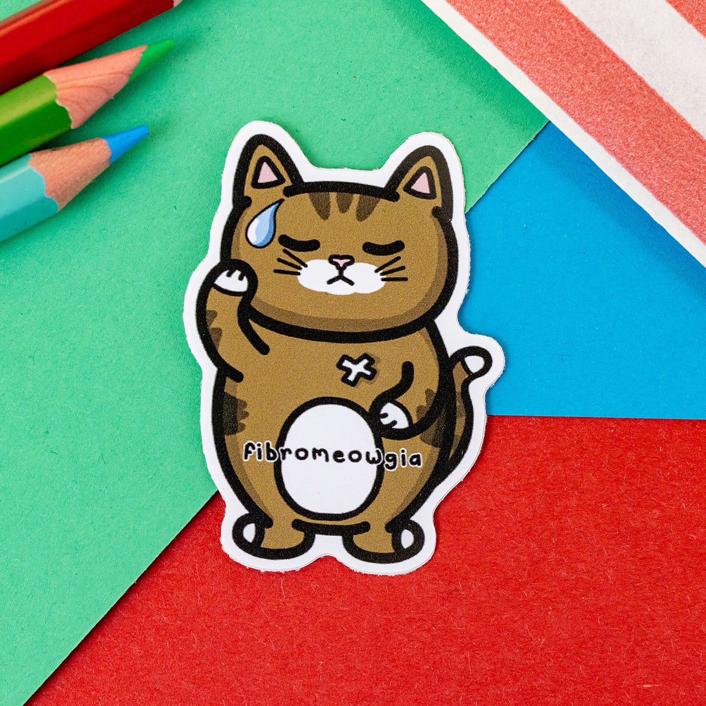 The Fibromeowgia Cat Sticker - Fibromyalgia Syndrome on a red, blue and green background with colouring pencils and a red stripe candy bag. The brown sad tabby cat shaped sticker has one paw on its head and the other on its tummy with a white bandaid and sweat droplet, across its middle reads 'fibromeowgia'. The hand drawn design is raising awareness for fibro fibromyalgia syndrome.