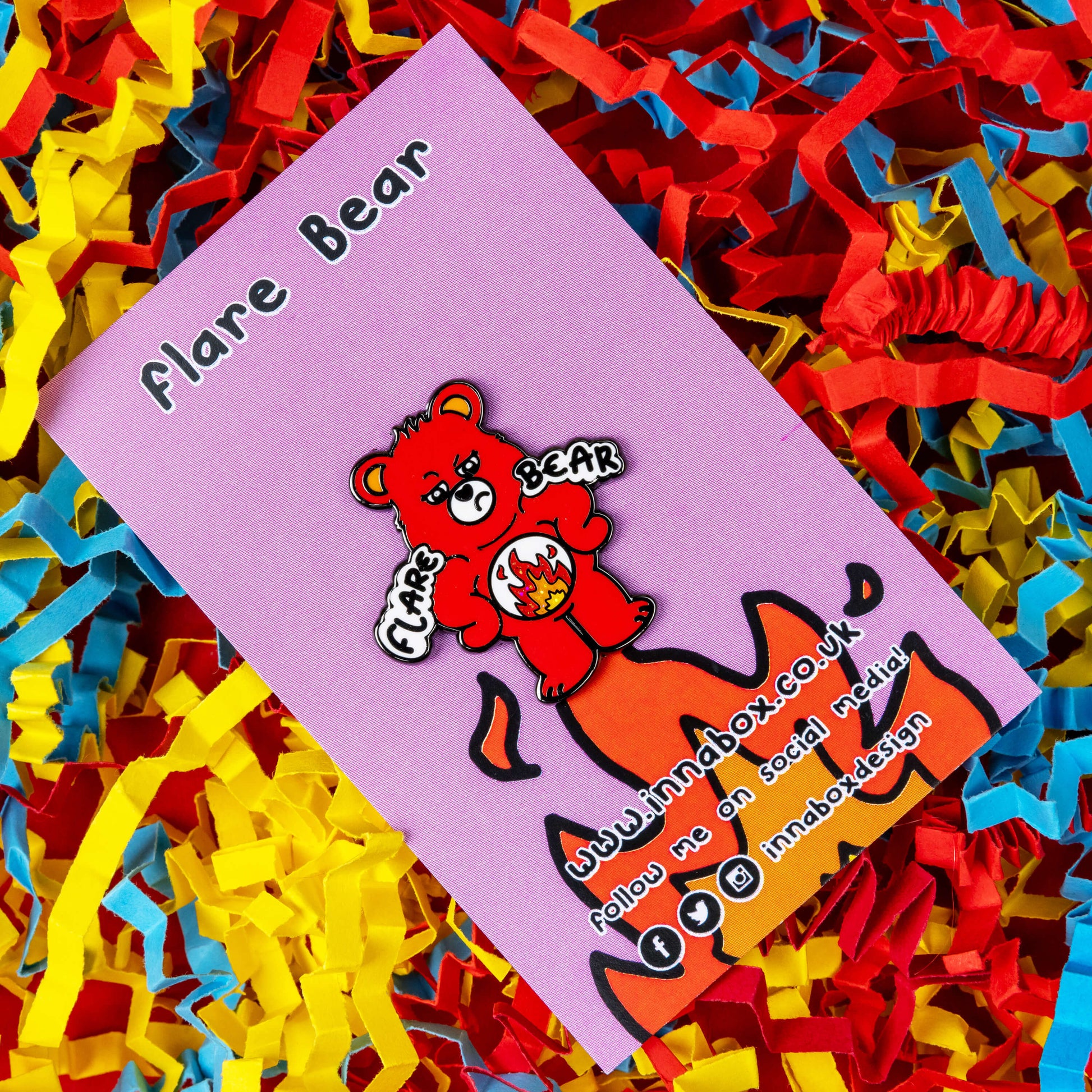 Flare Bear enamel pin on pink backing card with flames on held in front of a red, blue and yellow card confetti background. The enamel pin is of a red bear with a fed up expression and hands on its hips. There is a white circle on its belly with flames inside. Flare Bear is written on the pin. The enamel pin is designed to raise awareness for chronic illness flare ups.