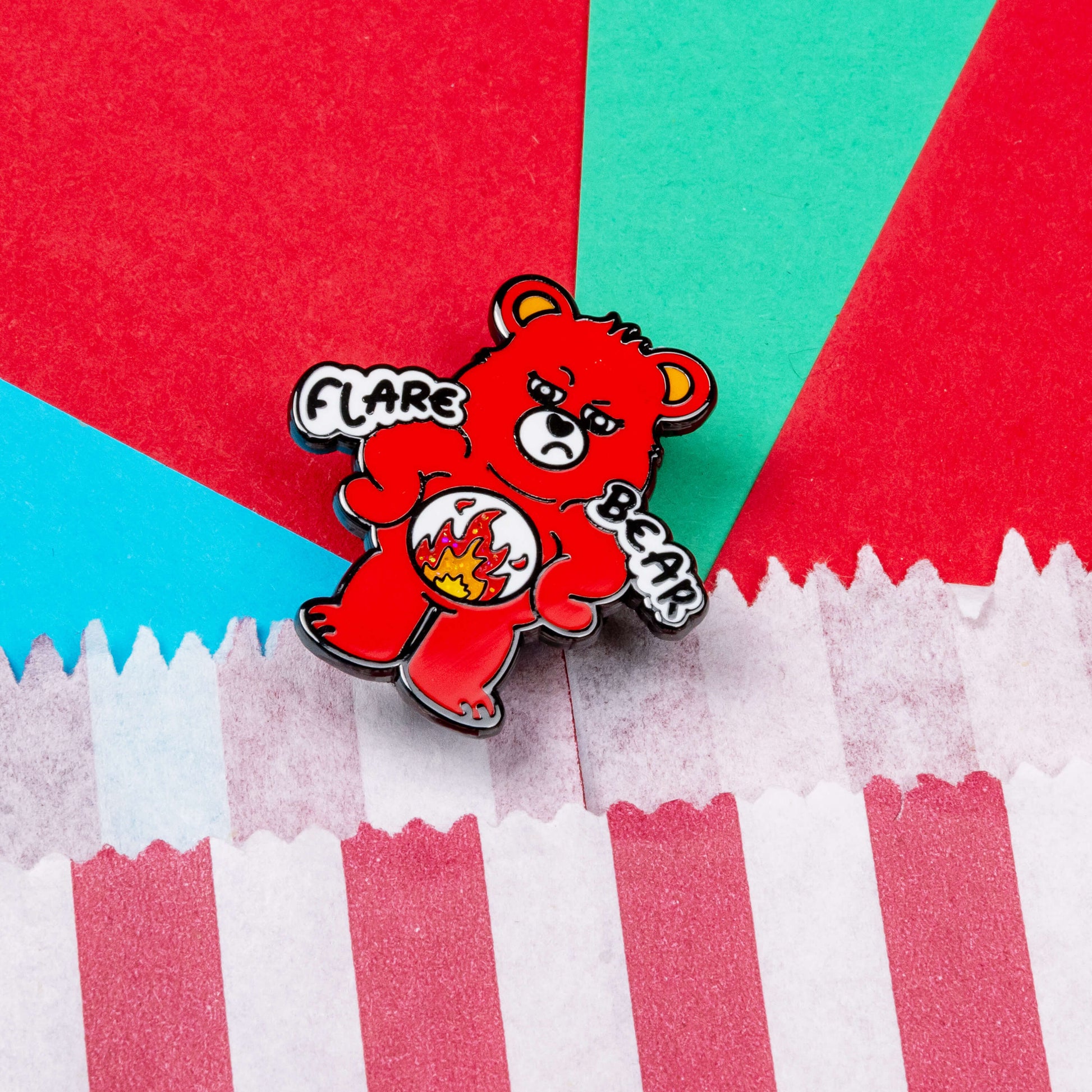 Flare Bear enamel pin shown on a red, blue and teal background next to a red and white striped paper bag. The enamel pin is of a red bear with a fed up expression and hands on its hips. There is a white circle on its belly with flames inside. Flare Bear is written on the pin. The enamel pin is designed to raise awareness for chronic illness flare ups.