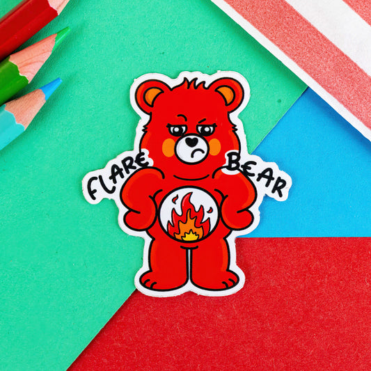 A sticker of a red bear looking grumpy with a flame on its tummy with the words FLARE BEAR on its shoulders it is on a green, red and blue background with pencils in in the corner of the shot. The hand drawn design is raising awareness for flare ups in chronic illnesses.