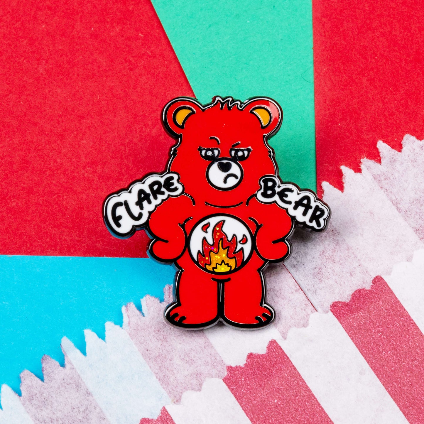 Flare Bear enamel pin shown on a red, blue and teal background next to a red and white striped paper bag. The enamel pin is of a red bear with a fed up expression and hands on its hips. There is a white circle on its belly with flames inside. Flare Bear is written on the pin. The enamel pin is designed to raise awareness for chronic illness flare ups.