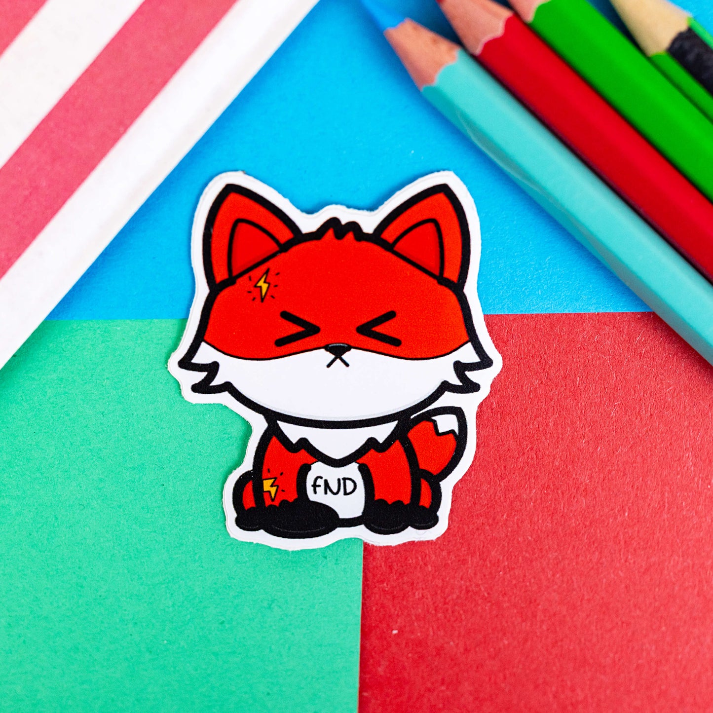 A red fox sticker looking very cute but in pain with the text FND on its stomach. It is on a red, blue and green background with coloured pencils to one side and a red stripy paper bag. The hand drawn design is raising awareness for Functional Neurological Disorder FND.