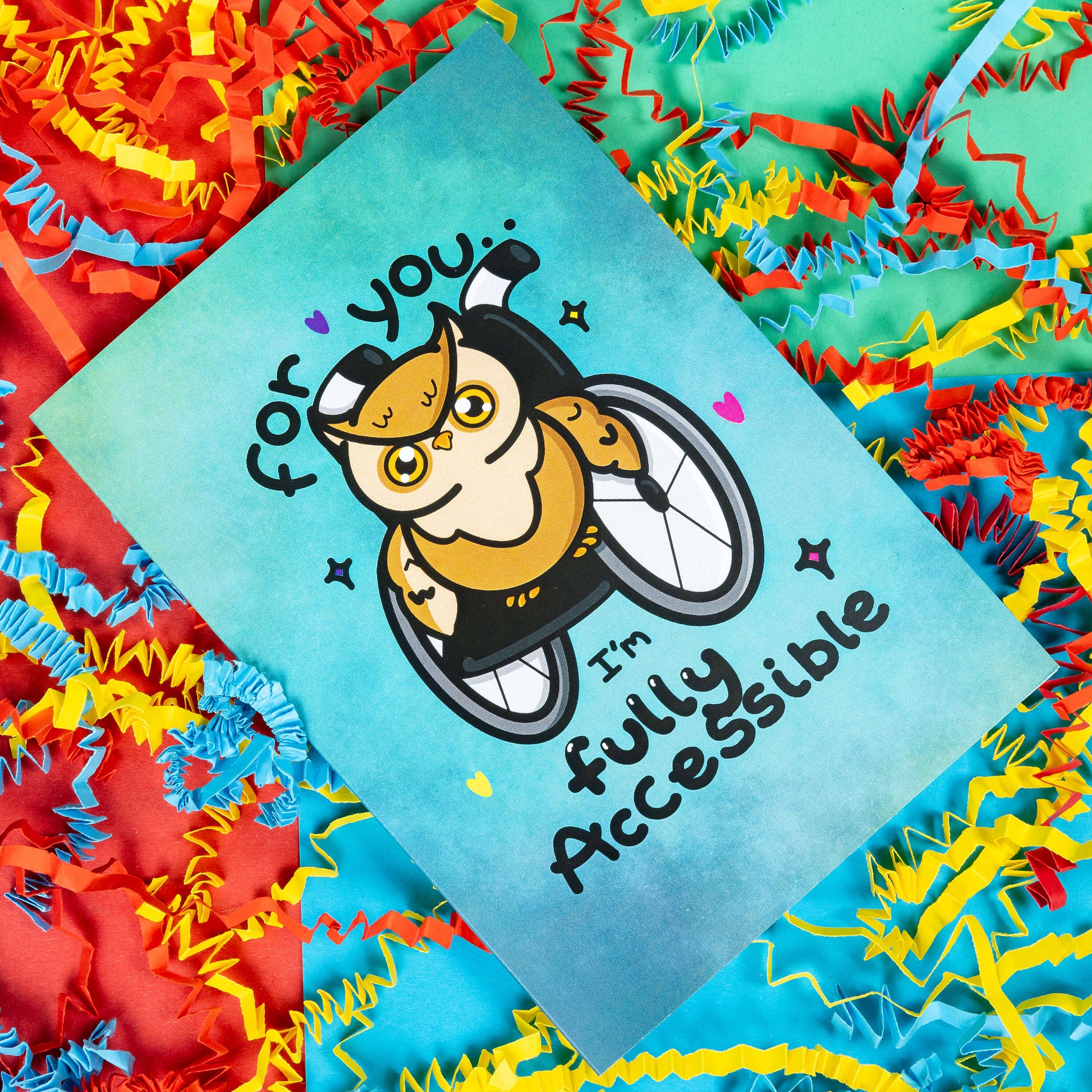 A blue greeting card with an illustration of a brown owl in a wheelchair with pink, purple and yellow love hearts and sparkles dotted around it. 'For you I'm fully accessible' is written on the card in black. The background of the photo is colourful card confetti. Design inspired by disabilities.