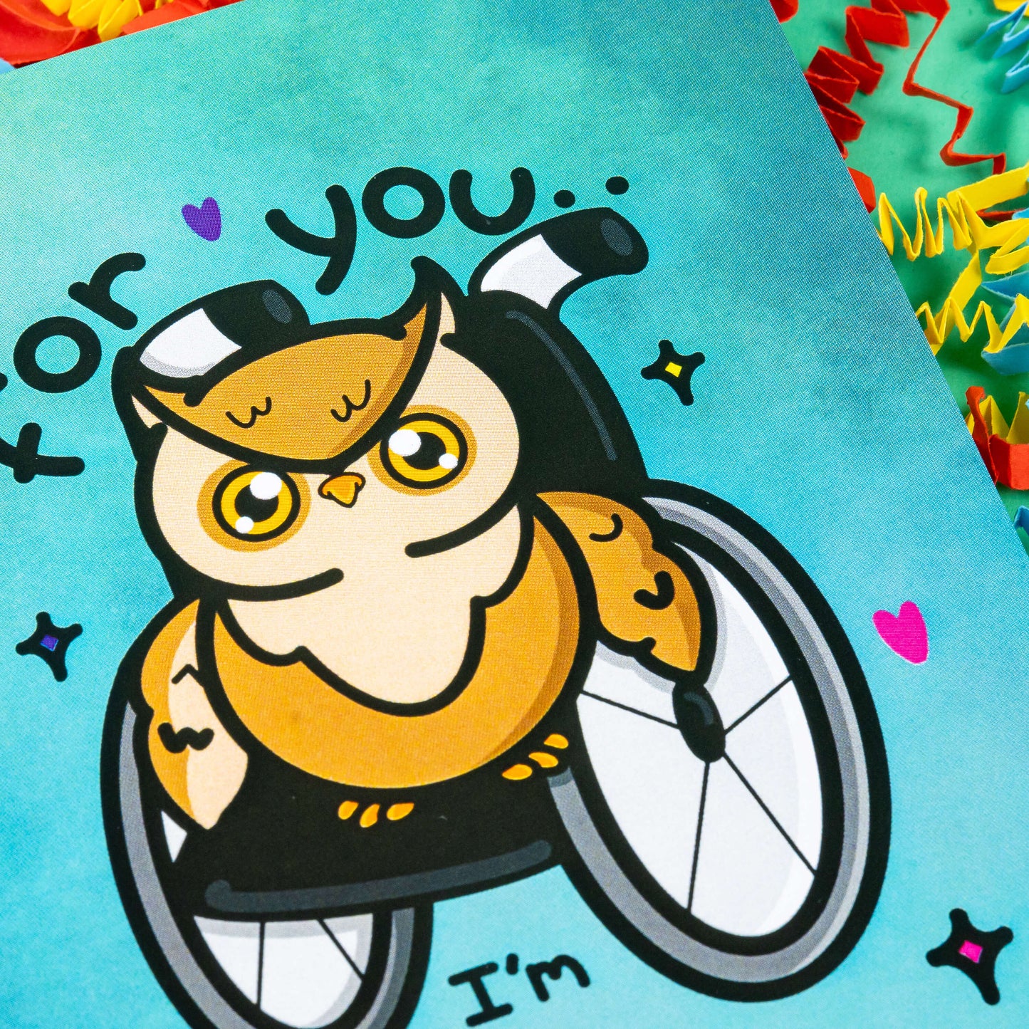 Close up of A blue greeting card with an illustration of a brown owl in a wheelchair with pink, purple and yellow love hearts and sparkles dotted around it. 'For you I'm fully accessible' is written on the card in black. The background of the photo is colourful card confetti. Design inspired by disabilities.