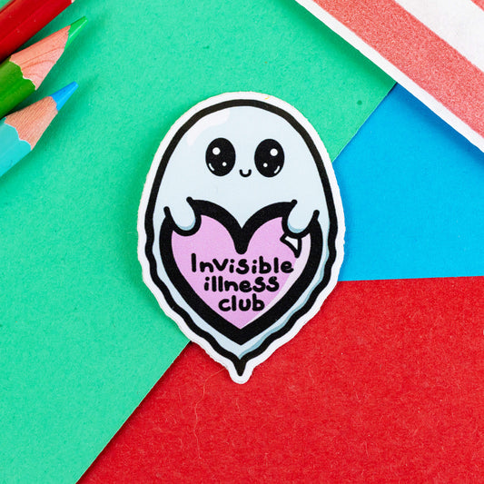 The Invisible Illness Club Sticker on a red, blue and green background with colouring pencils and red stripe candy bag. The pastel blue smiling ghost sticker has big sparkly eyes holding a pastel pink heart with black text reading 'invisible illness club'. The hand drawn design is raising awareness for hidden disabilities.