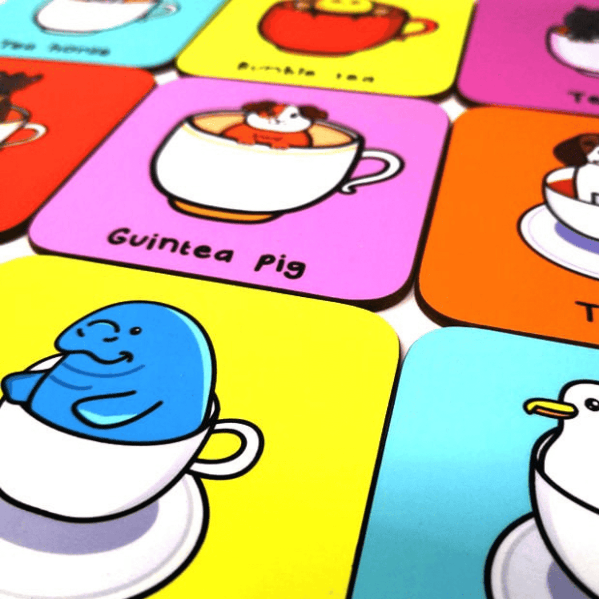 The Guinea Pig Tea Coaster amongst other innabox tea pun coasters. The pastel pink wooden coaster has a smiling brown and white guinea pig popping out of a white mug of tea, underneath in black reads 'guintea pig'.