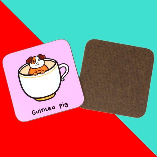The Guinea Pig Tea Coaster on a red and blue background. The pastel pink wooden coaster has a smiling brown and white guinea pig popping out of a white mug of tea, underneath in black reads 'guintea pig', to the right is the dark brown wooden underside of the coaster.