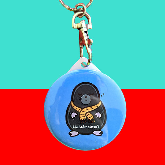 Hashimoto's Thyroiditis - Mole Keyring on a red and blue background. The blue circular keyring has an illustration of a sleeping mole wearing a yellow scarf. The mole has white text across his tummy that reads 'hashimoleto's'. Hand drawn design is made to raise awareness for Hashimoto's Thyroiditis and autoimmune diseases.