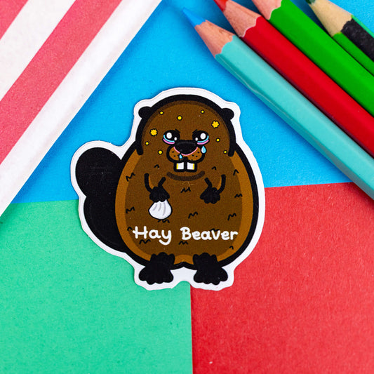 Hay Beaver Sticker on a red, blue and green background with colouring pencils and red stripe candy bag. The brown beaver sticker has watery eyes, dripping nose and yellow spots holding a tissue with 'hay beaver' written in white across its lower body. The hand drawn design is raising awareness for hay fever and allergic rhinitis.