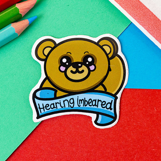 Hearing Imbeared Bear Sticker - Hearing Impaired on a red, green and blue background with colouring pencils. The cute brown bear sticker with big sparkly eyes and rosy cheeks holding one hand to its ear. There is a blue banner under the bear with 'Hearing imbeared' written across in black writing. The hand drawn sticker is raising awareness for hearing impaired people.