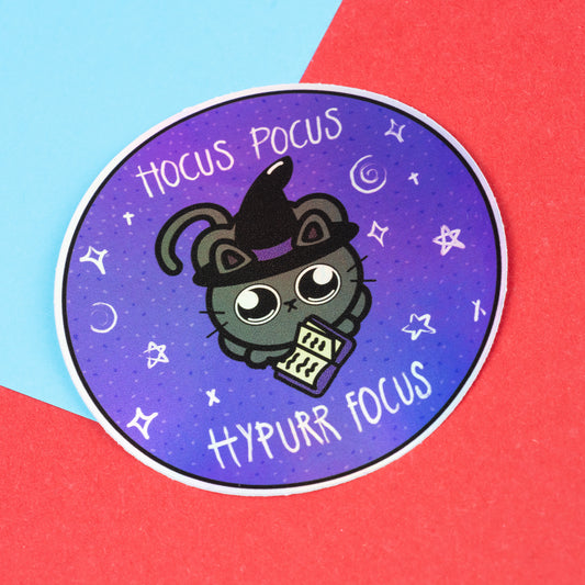 A holographic vinyl sticker by Innabox featuring a cute black cat wearing a witch hat, holding a spell book. Surrounded by stars and moons, it reads “Hocus Pocus Hypurr Focus.” Set on a red and blue paper background. Ideal for those with ADHD or chronic illness.