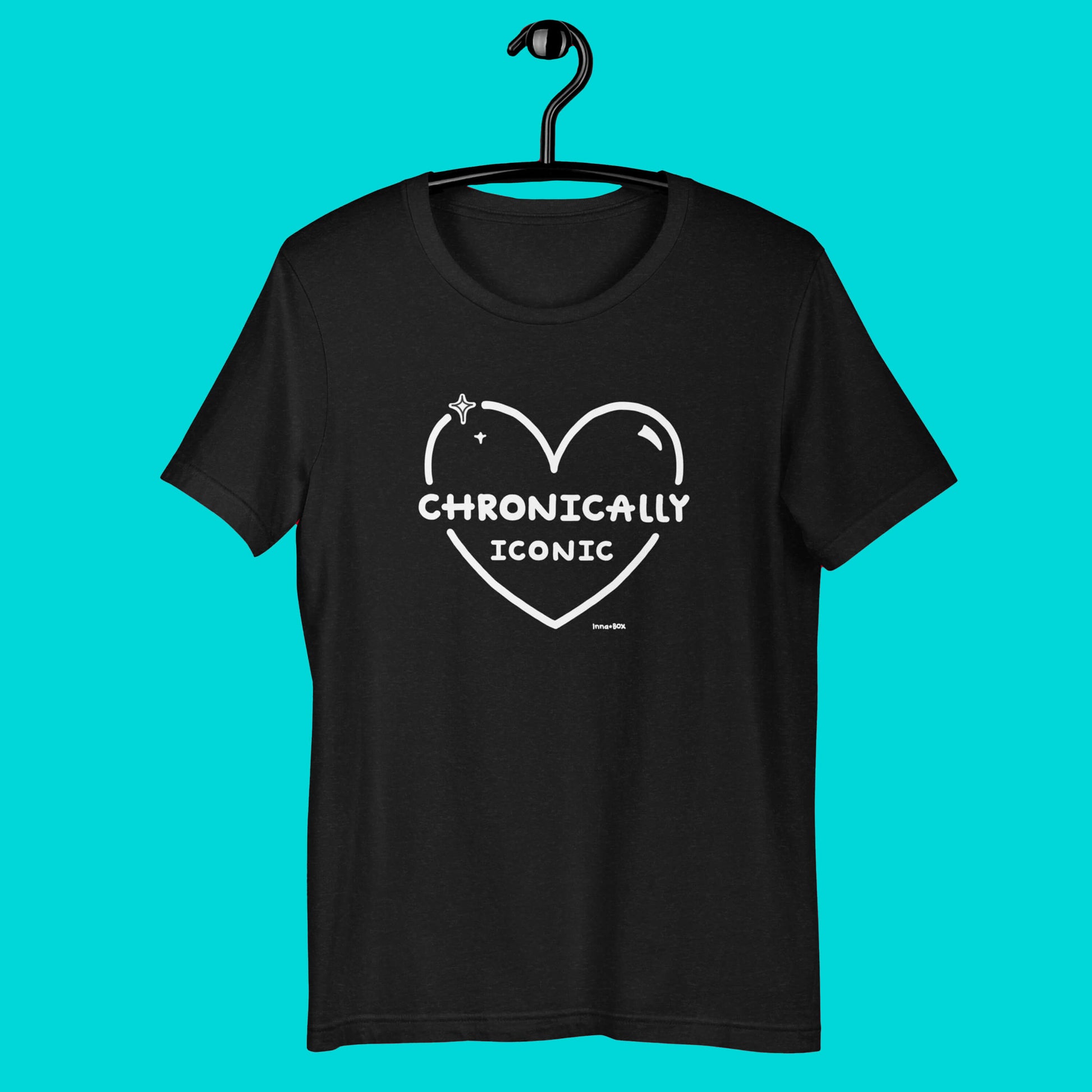 The Chronically Iconic black tee hanging up on a black hanger over a blue background. The short sleeve t-shirt features a white heart outline with sparkles and centre text reading 'chronically iconic' with the innabox logo underneath. The design is raising awareness for chronic illness and invisible illness.