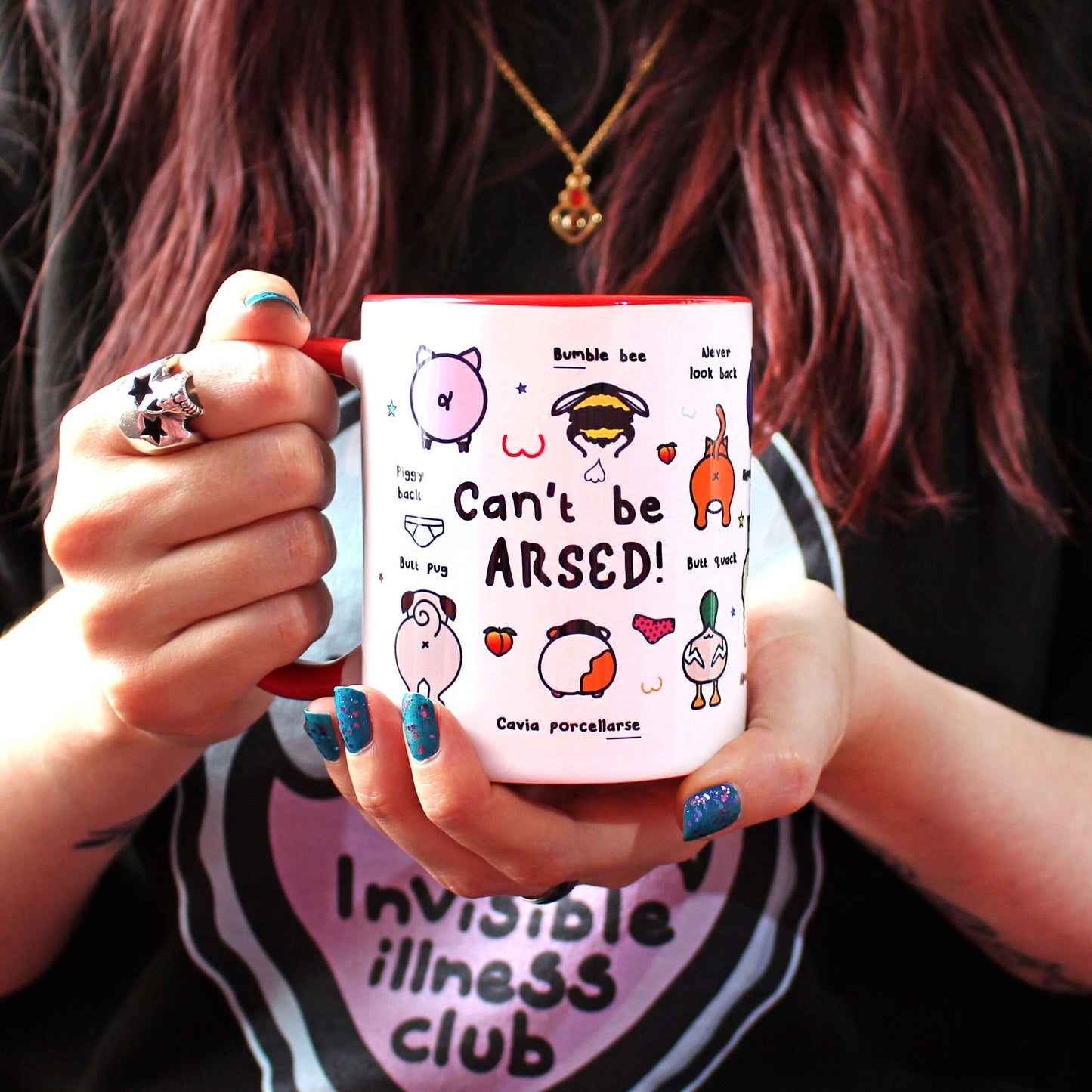 The Can't Be Arsed Mug being held up by Nikky Box with brown hair and blue nail varnish wearing the invisible illness black sweater. The white mug with a red handle and inside features various animal bums with underwear, chest outlines, peaches and multicoloured stars. The mug is facing right which shows pig - piggy back, bumble bee - bum ble bee, pug - butt pug, guinea pig - cavia porcellarse, duck - butt quack and cat - never look back, bums.
