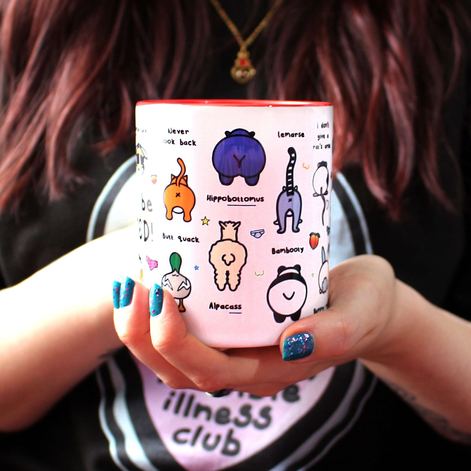 The Can't Be Arsed Mug being held up by Nikky Box with brown hair and blue nail varnish wearing the invisible illness black sweater. The white mug with a red handle and inside features various animal bums with underwear, chest outlines, peaches and multicoloured stars. The mug is facing forward which shows bumble bee - bum ble bee, guinea pig - cavia porcellarse, duck - butt quack, cat - never look back, hippo - hippobottomus, alpaca - alpacass, panda - bambooty and lemur - lemarse, bums.