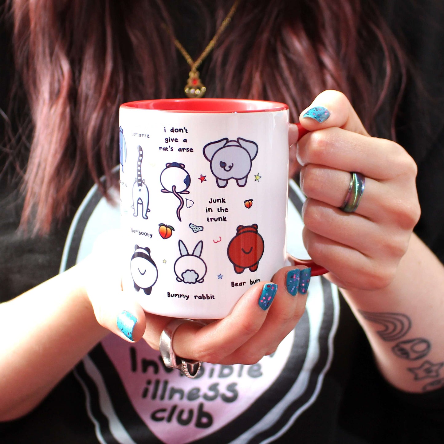 The Can't Be Arsed Mug being held up by Nikky Box with brown hair and blue nail varnish wearing the invisible illness black sweater. The white mug with a red handle and inside features various animal bums with underwear, chest outlines, peaches and multicoloured stars. The mug is facing left which shows hippo - hippobottomus, alpaca - alpacass, panda - bambooty, lemur - lemarse, rat - I don't give a rat's arse, bunny rabbit - bummy rabbit, elephant - junk in the trunk and bear - bear bum bums.