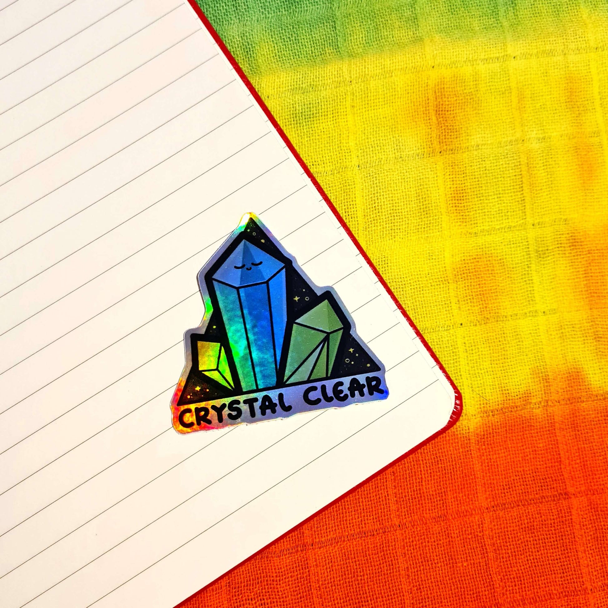 The Crystal Clear Holographic Sticker stuck inside a lined notebook on a rainbow tie dye fabric. The triangle shaped sticker features three green crystal towers on a black sparkly background with bottom text reading crystal clear, the centre crystal is smiling. Inspired by witchy spiritual healing with crystals.