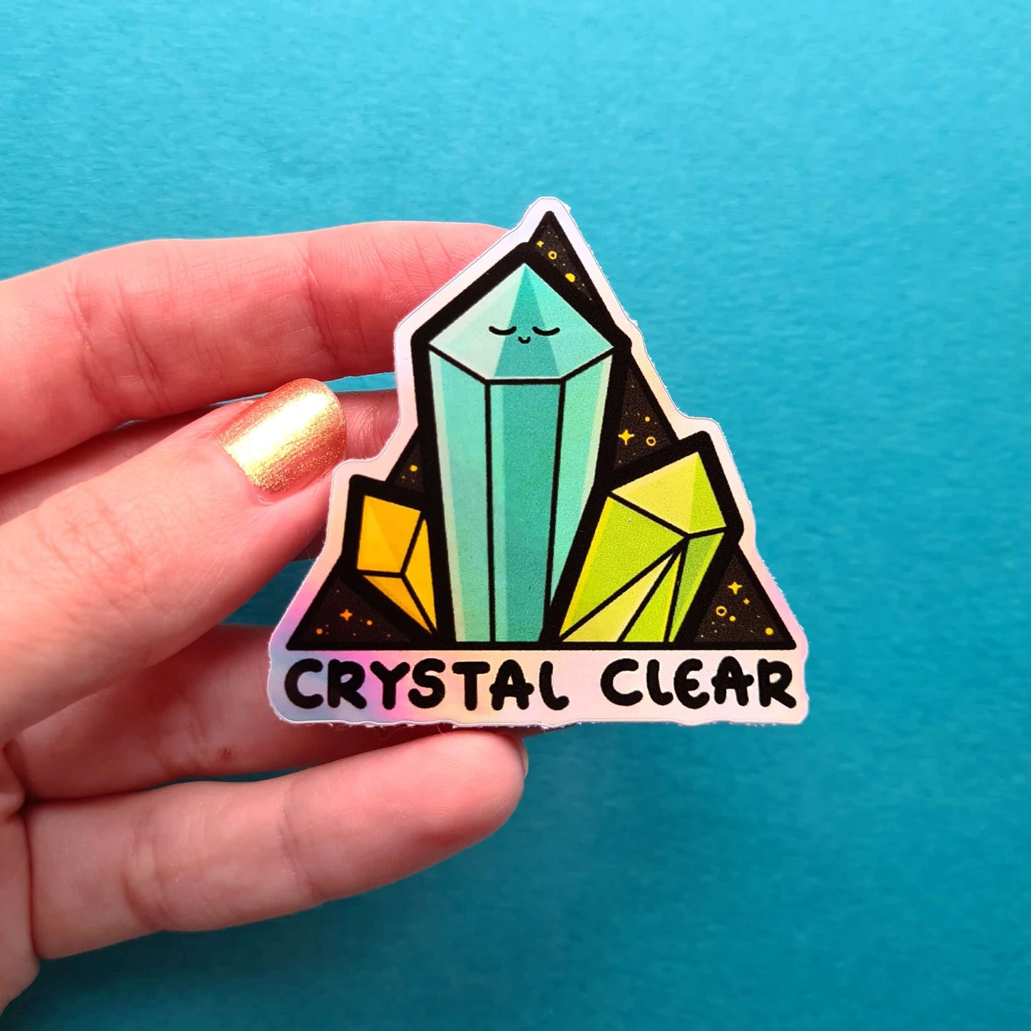 The Crystal Clear Holographic Sticker being held over a blue background. The triangle shaped sticker features three green crystal towers on a black sparkly background with bottom text reading crystal clear, the centre crystal is smiling. Inspired by witchy spiritual healing with crystals.