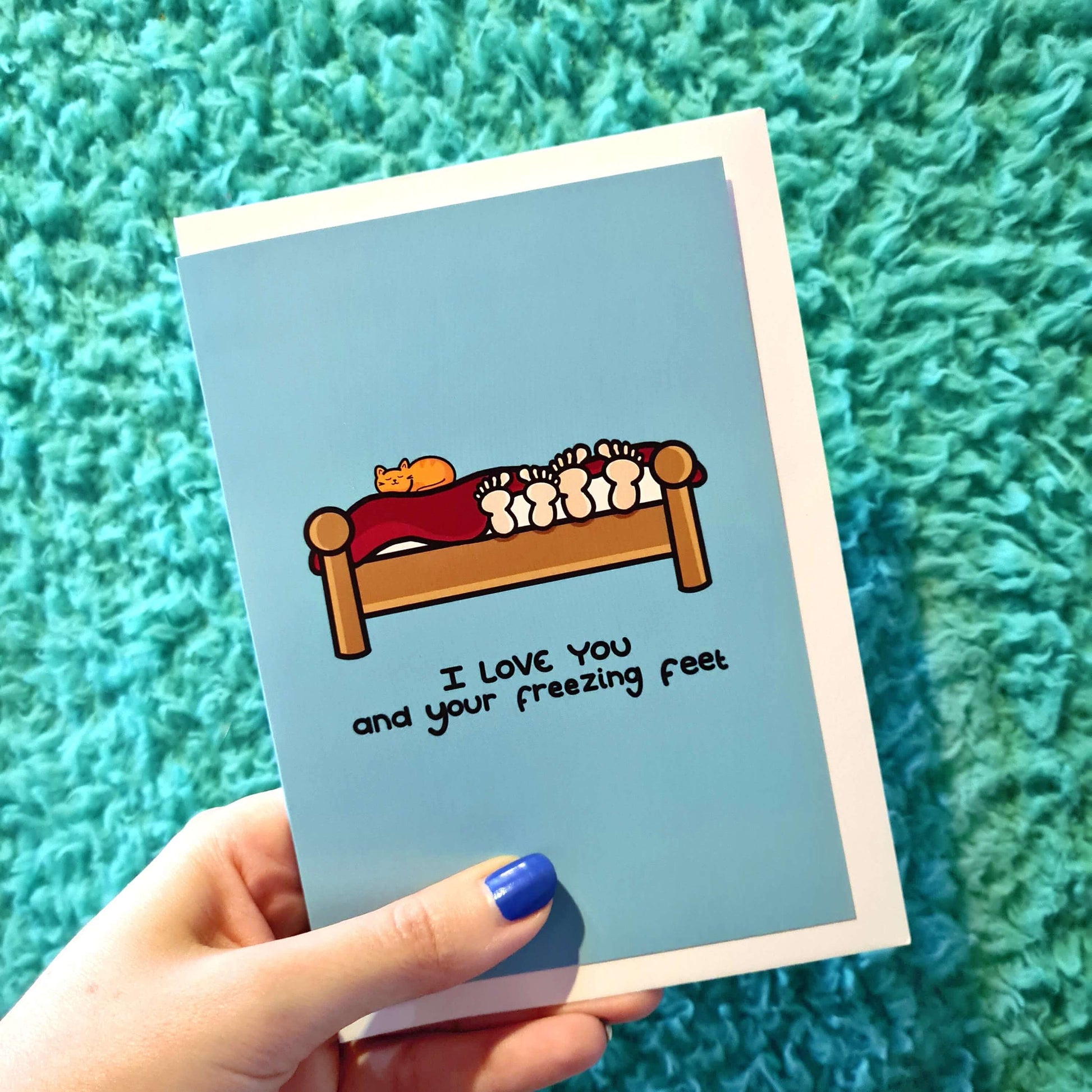 I love You And Your Freezing Feet Card held in front of a blue blanket. the light blue card has an illustration of a wooden bed with two pairs of feet poking out from under the red blanket and a ginger cat sat on top of it