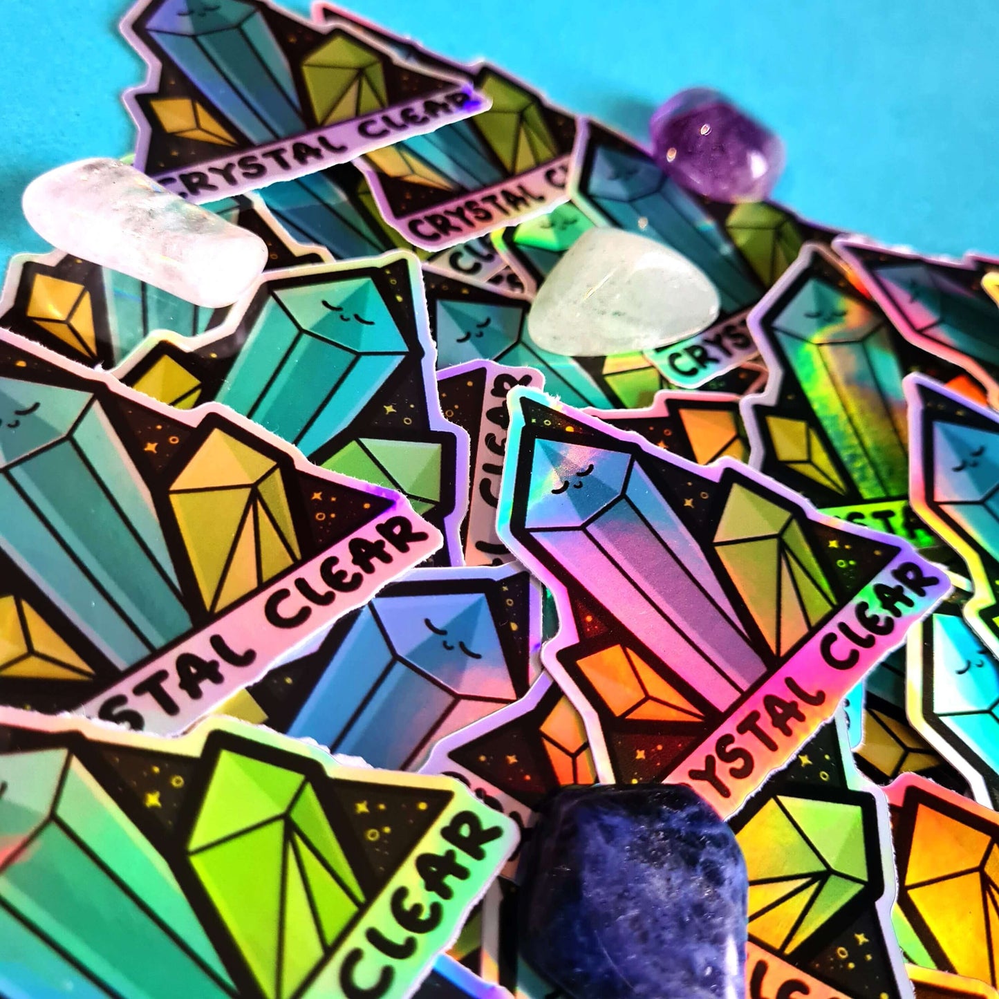 Crystal Clear Holographic Stickers pushed into a triangle shape with various crystals (amethyst, clear quartz, lapis lazuli, jasper) on a blue background. The triangle shaped sticker features three green crystal towers on a black sparkly background with bottom text reading crystal clear, the centre crystal is smiling. Inspired by witchy spiritual healing with crystals.