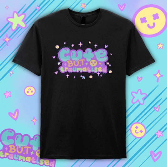 Black “Cute But Traumatised” T-shirt by Innabox featuring colourful pastel typography with stars, hearts and smiley details. Soft, playful graphic design contrasts with a meaningful message supporting mental health and hidden disabilities. Unisex cotton tee laid flat against a bright illustrated background, ideal for alternative fashion lovers.