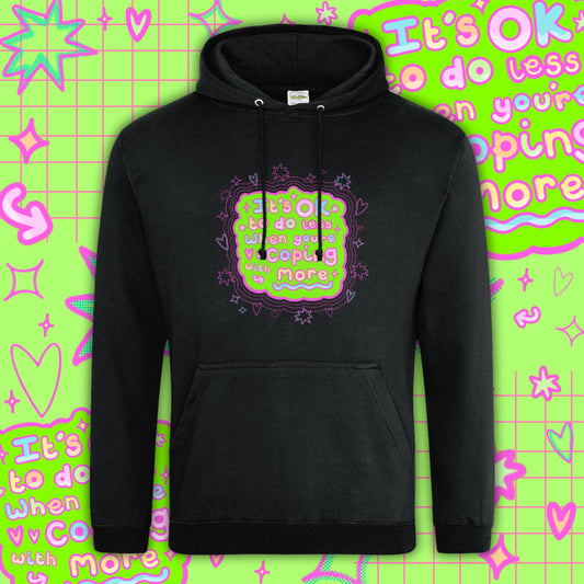 Black hoodie featuring a colourful neon graphic reading “It’s OK to do less when you’re coping with more” by Innabox, a disability awareness brand. The hoodie has a front pocket, drawstring hood and soft cotton fabric, designed for comfort and mental health support.