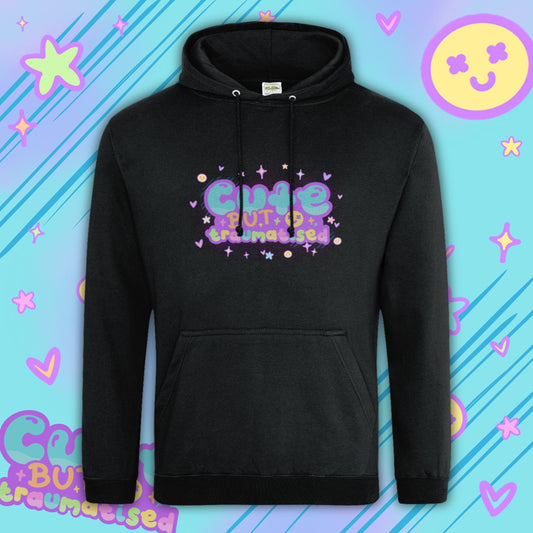 Black hoodie by Innabox featuring colourful “Cute But Traumatised” slogan in pastel purple, green and yellow bubble lettering. Soft cotton blend hoodie with front pouch pocket, drawstring hood and relaxed fit. Disability awareness clothing designed to uplift people with chronic illness and hidden disabilities.
