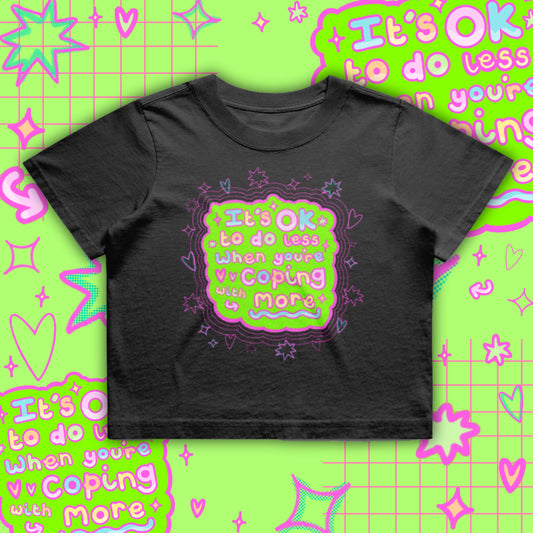 Black cropped t-shirt by Innabox featuring a bright neon green and pink graphic reading “It’s OK to do less when you’re coping with more”. Boxy oversized fit with a soft cotton finish. Disability awareness clothing designed to support people with chronic illness, mental health struggles and hidden disabilities.