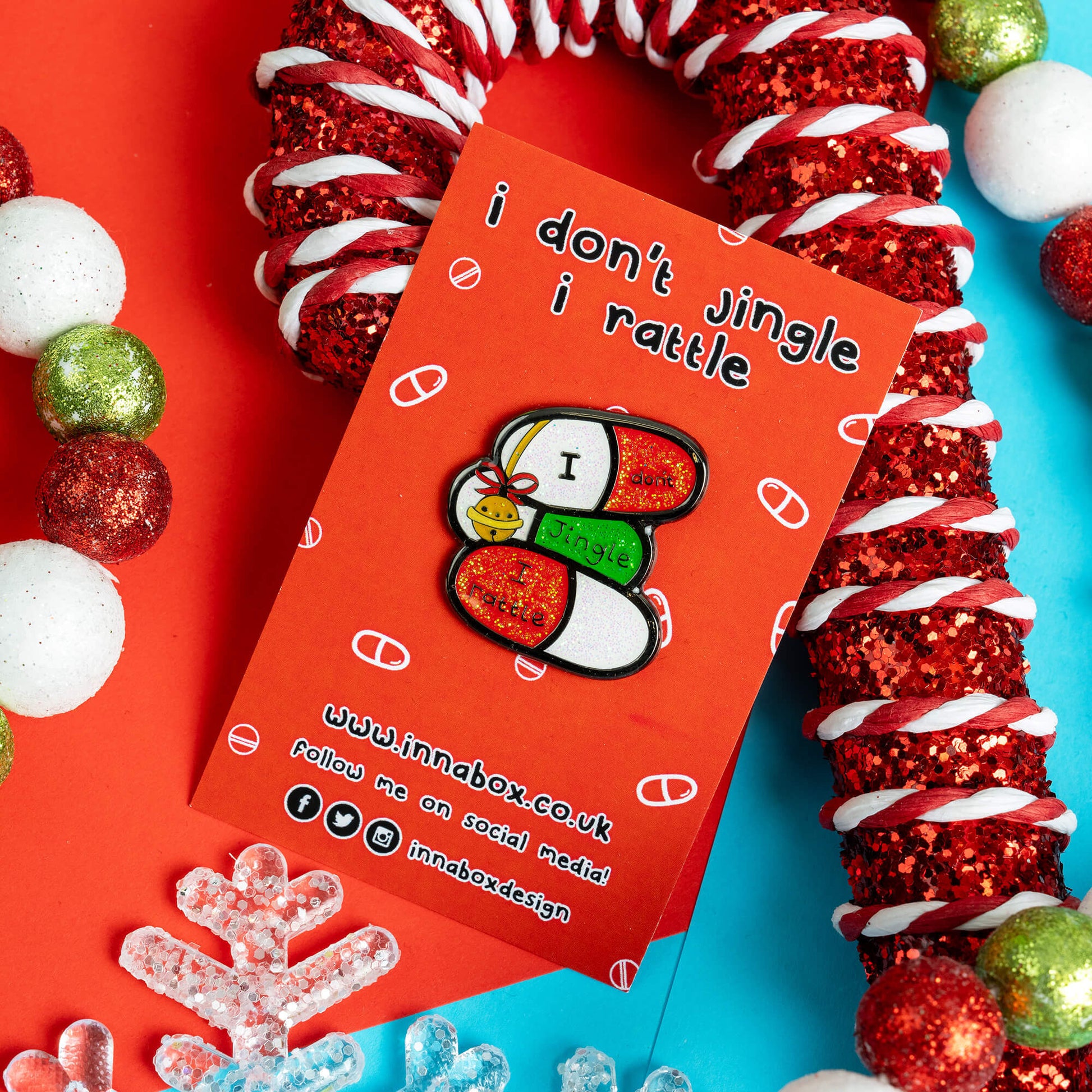 Innabox enamel pin featuring three pill capsules with the phrase 'I don’t jingle, I rattle' on a red backing card with the same phrase. It's laid on a vibrant festive background of red and blue with snowflake decorations. A playful and empowering design for people with chronic illnesses or hidden disabilities.