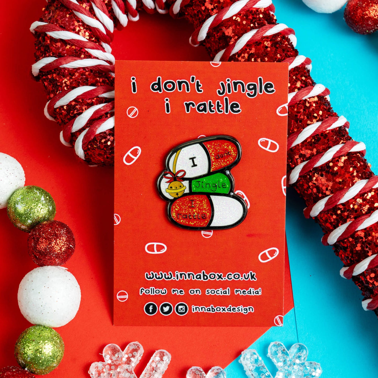 Innabox enamel pin featuring three pill capsules with the phrase 'I don’t jingle, I rattle' on a red backing card with the same phrase. It's laid on a vibrant festive background of red and blue with snowflake decorations. A playful and empowering design for people with chronic illnesses or hidden disabilities.