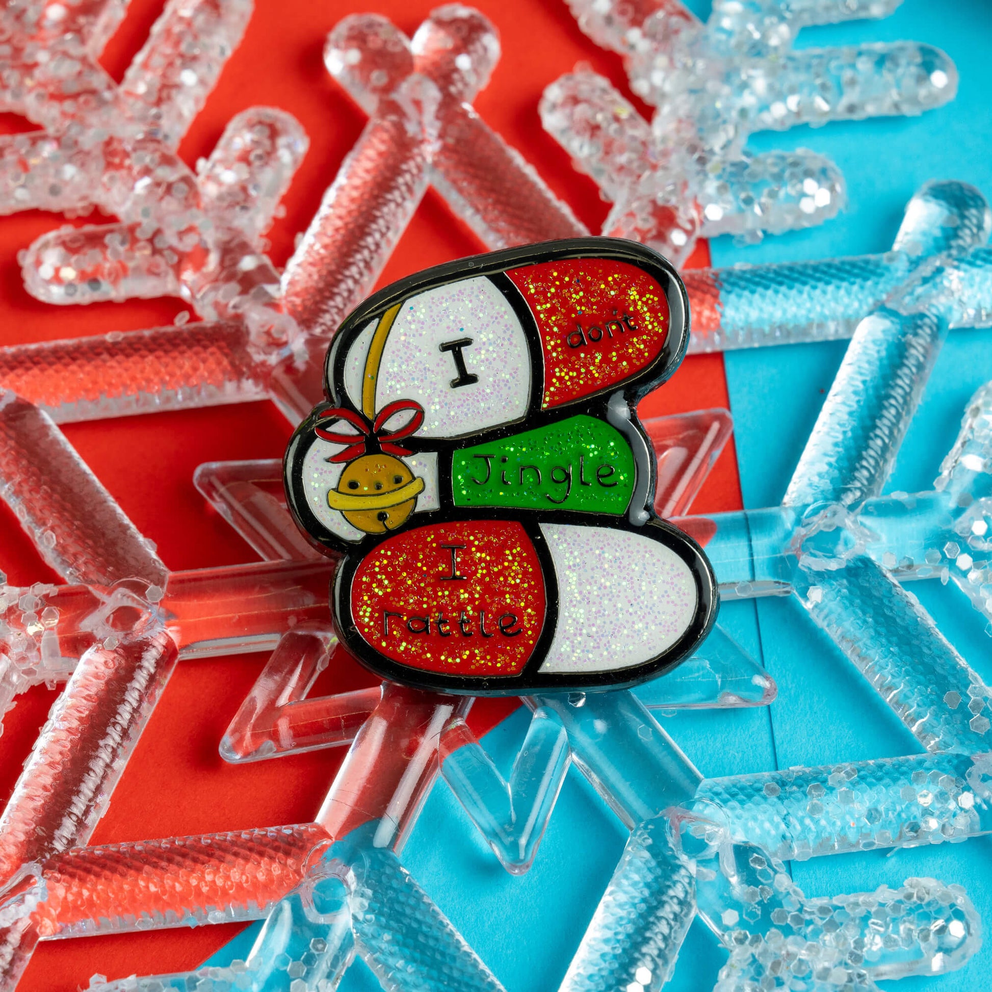 Innabox enamel pin featuring three capsules with the phrase 'I don’t jingle, I rattle' against a vibrant background of red and blue with snowflake decorations. A playful and empowering design for people with chronic illnesses or hidden disabilities.