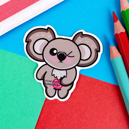The Ulcerative Koalitis Koala Sticker - Ulcerative Colitis UC on a red, blue and green background with colouring pencils and red stripe candy bag. The grey koala shaped vinyl sticker has one eye shut clutching its red swirly tummy with black text over it reading 'Ulcerative Koalitis'. The hand drawn design is raising awareness for Ulcerative Colitis UC.