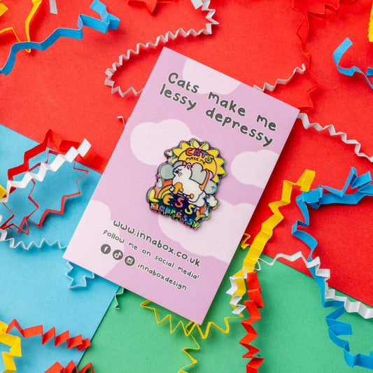 An Innabox enamel pin displayed on its branded backing card. The pin features a cat, rainbow, and sun, with the phrase “Cats make me lessy depressy.” The colourful background includes red, green, and blue sections with curly paper decorations, enhancing the vibrant look. Innabox creates products to support those living with hidden disabilities and chronic illnesses.