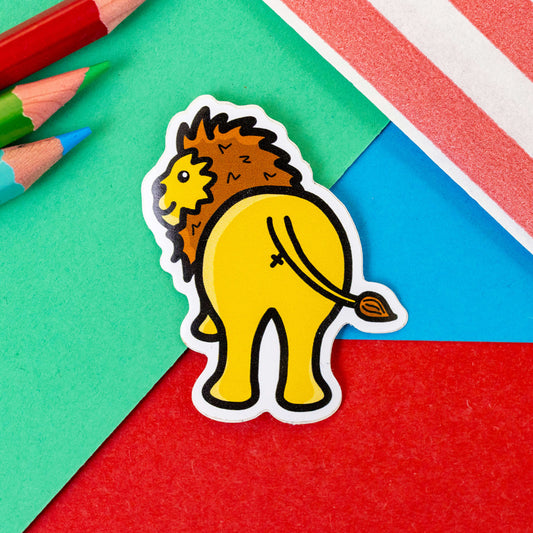 The Lion Bum Sticker on a red, blue and green background with colouring pencils and red stripe candy bag. The yellow lion with brown mane is facing away showing its bum smiling looking over its shoulder. A cheeky gift or decoration.