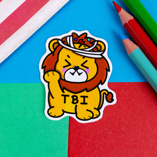 Traumatic Brain Injury sticker on a red and blue background. The sticker is in the shape of a cute lion which is orange with a brown mane and tip of tail. The lion's eyes are screwed up shut with rose cheeky and two little teeth poking out of it's mouth. It has one paw holding its face. The lion has white bandages on its head with multi coloured dots around it. 'TBI' is black letters on the lion's stomach.