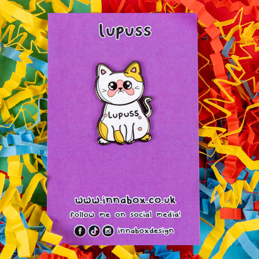 Lupuss Cat Enamel Pin - Lupus on it's purple backing card on a red, blue and yellow card confetti background. The cat shape enamel pin is orange and white with a sad expression, pink patches all over and black text across its middle reading 'lupuss'. The hand drawn design is raising awareness for lupus.