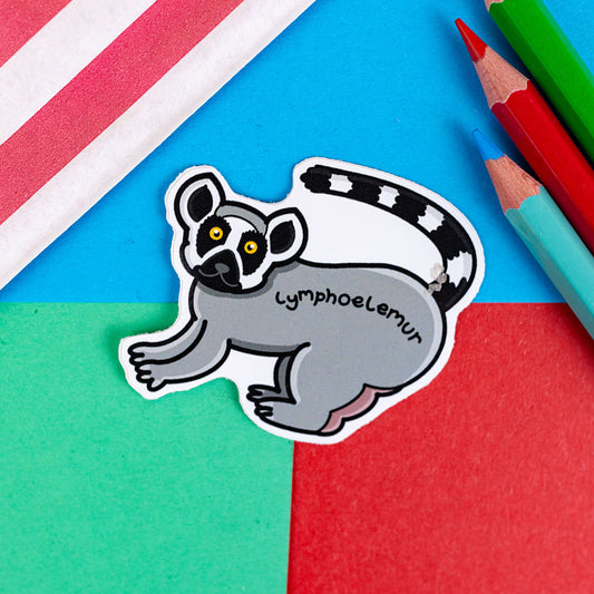Lymphoedema sticker shown on a red and blue background. The sticker is of a lemur standing on all fours and head turned to the side to that it is straight on. The lemur has yellow eyes and a black and white tail which is in the air. 'Lymphoelemur' is written across the body in black writing. It is on a red, blue and green background with coloured pencils to one side and a stripy paper bag