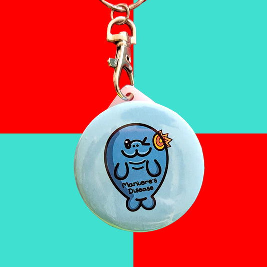 The Maniere's Disease Manatee Keyring - Ménière's Disease on a red and blue background. The pastel blue circular plastic keychain with silver lobster clip features a winking manatee with a yellow and red inflamed ear and black text across its body reading 'maniere's disease'. The hand drawn design is raising awareness for Ménière's Disease and vertigo.