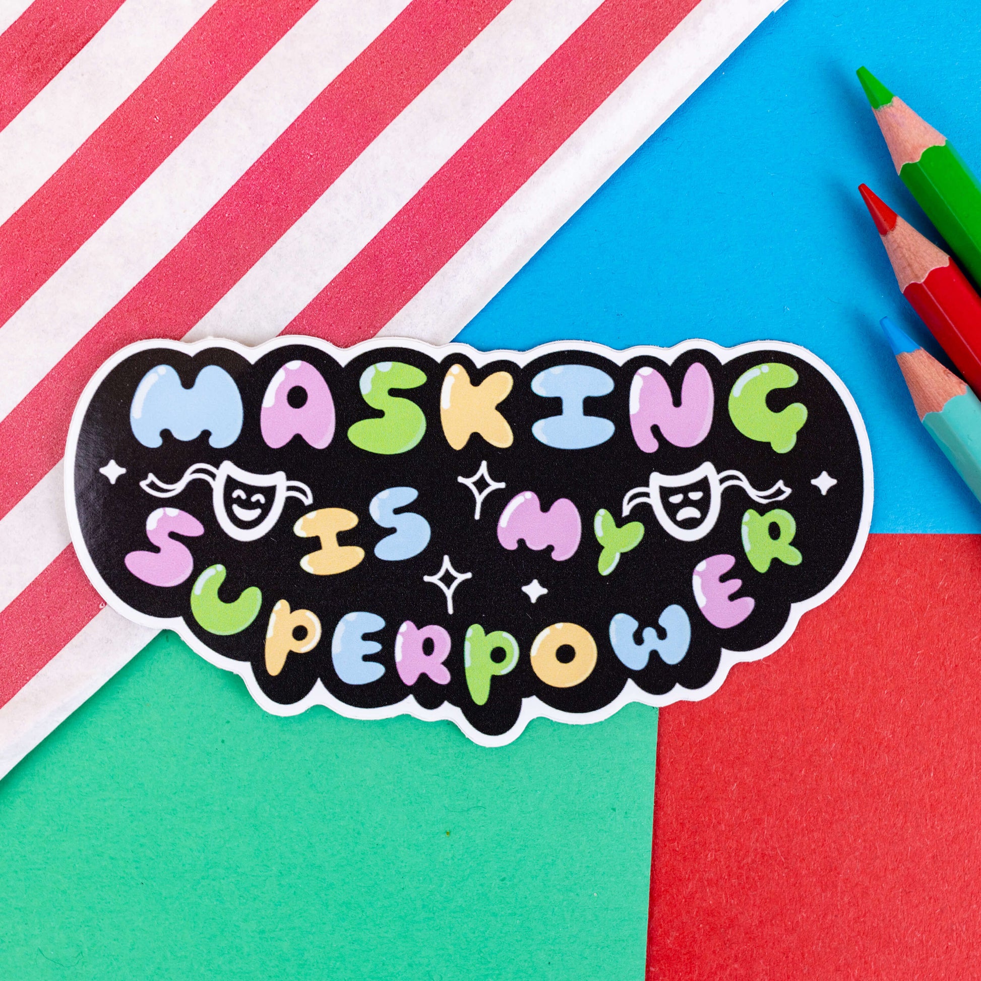 The Masking is my Superpower Sticker on a red, blue and green background with a red stripy candy bag and colouring pencils. The black base sticker features pastel rainbow bubble writing that reads 'masking is my superpower' with white sparkles, a happy and sad drama masks all on a black oval. Raising awareness for neurodivergent people with ADHD or Autism.