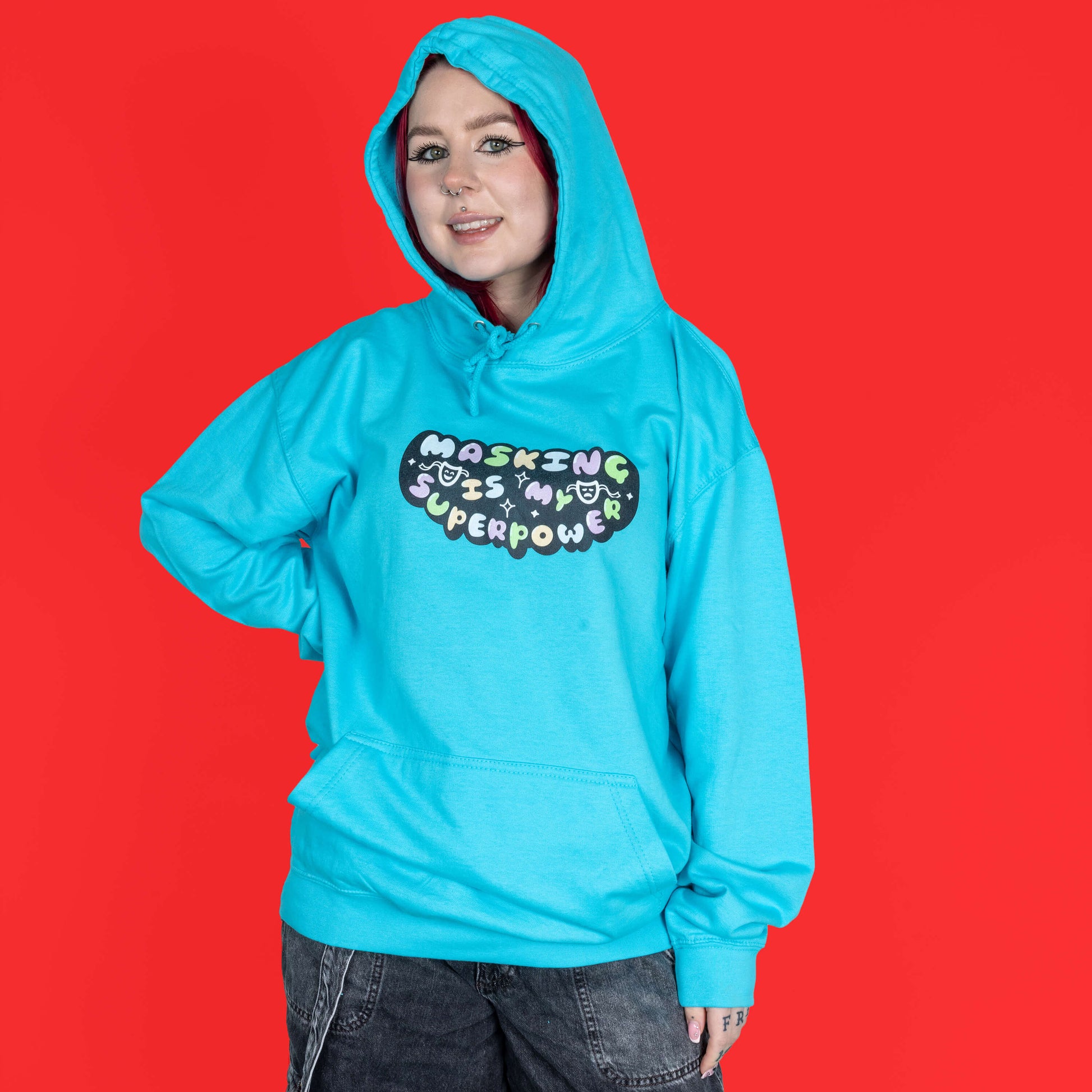 The Masking Is My Super Power Hoodie in Turquoise Surf modelled by Flo with red hair and black eyeliner on a red background. She is facing forward smiling with the hood up and one hand on her hip. The aqua blue hoodie features pastel rainbow bubble writing that reads 'masking is my superpower' with white sparkles, a happy and sad drama masks all on a black oval. Raising awareness for neurodivergent people with ADHD or Autism.
