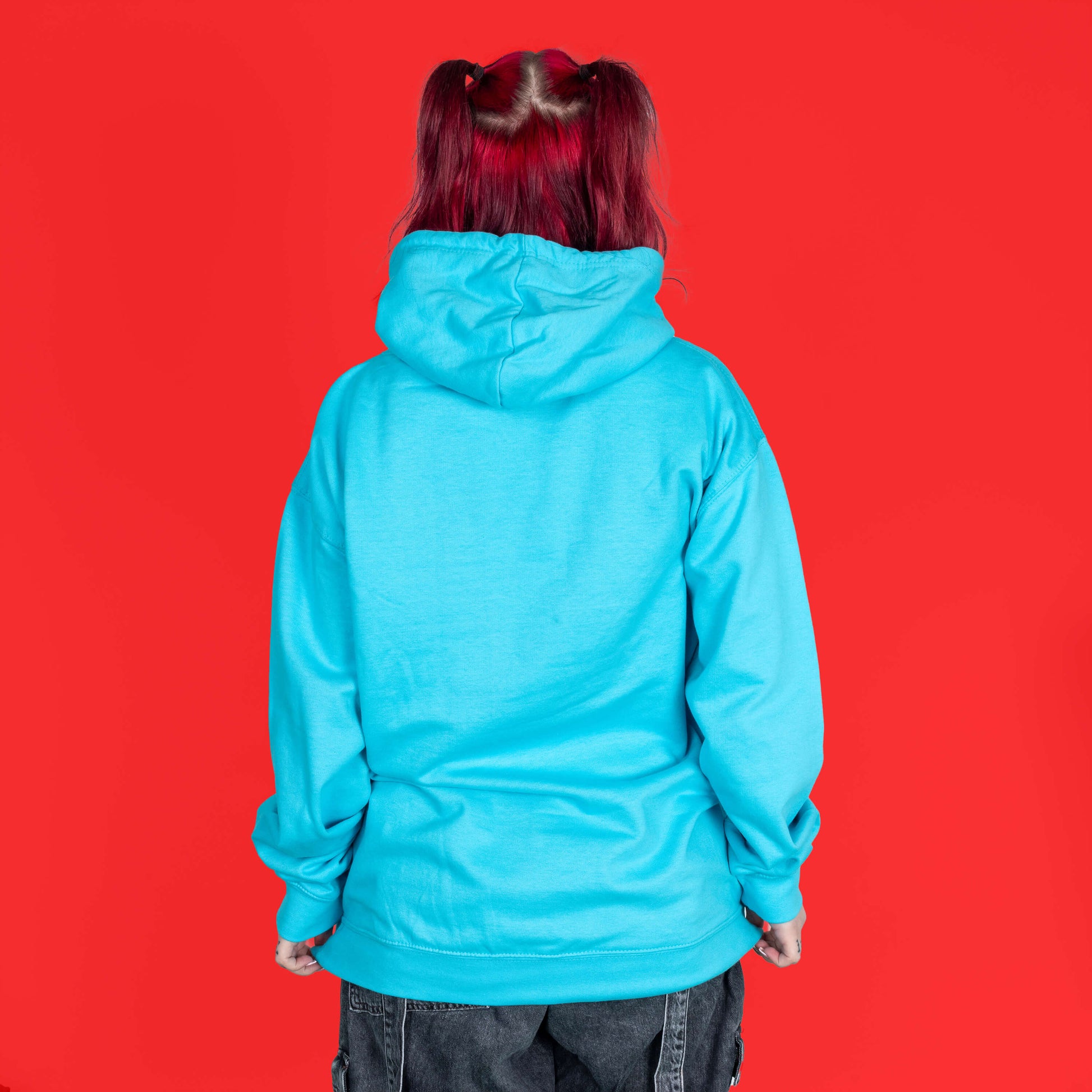 The Masking Is My Super Power Hoodie in Turquoise Surf modelled by Flo with red hair on a red background. She is facing away to highlight the plain back of the hoodie. The front of the aqua blue hoodie features pastel rainbow bubble writing that reads 'masking is my superpower' with white sparkles, a happy and sad drama masks all on a black oval. Raising awareness for neurodivergent people with ADHD or Autism.