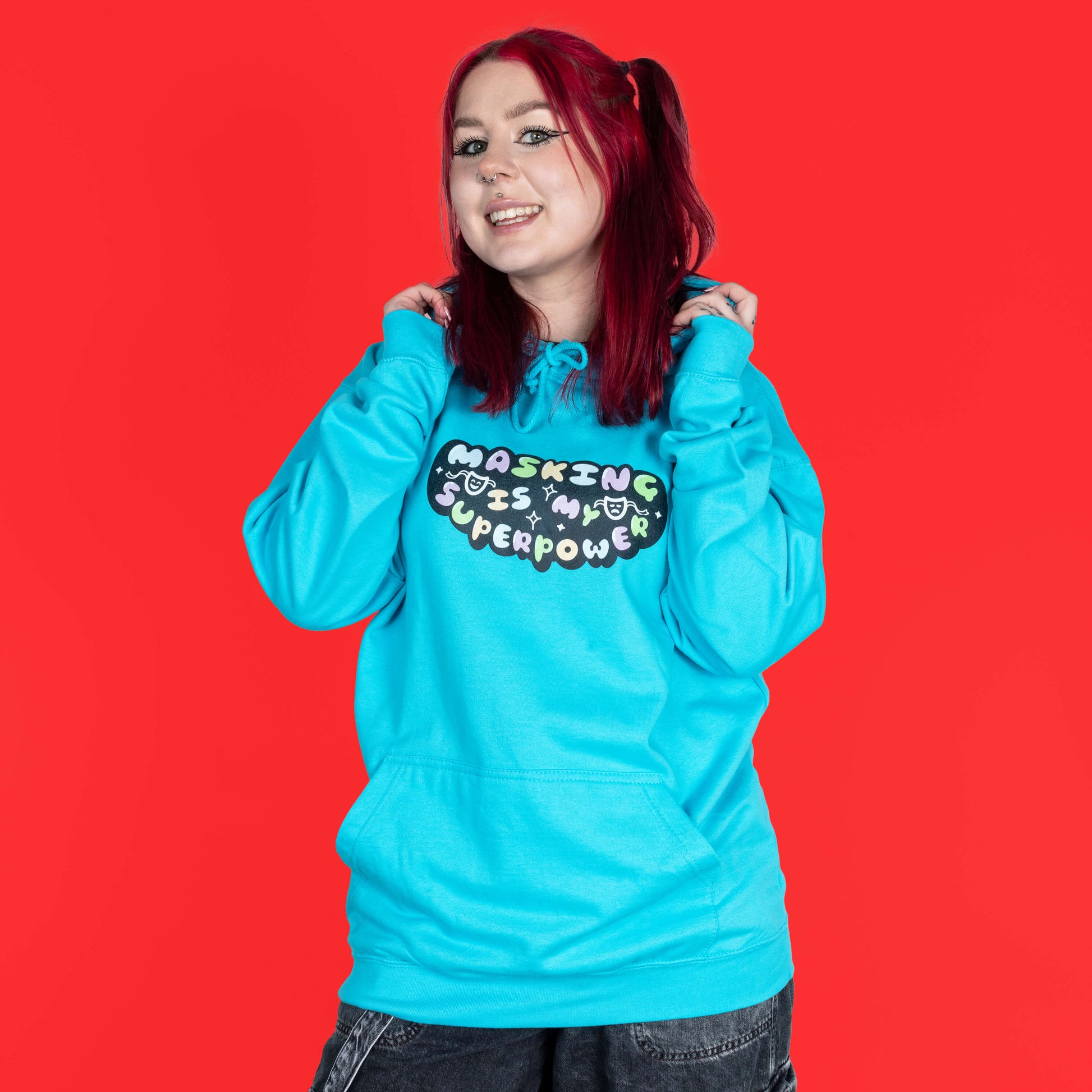 The Masking Is My Super Power Hoodie in Turquoise Surf modelled by Flo with red hair and black eyeliner on a red background. She is facing forward smiling holding the hood. The aqua blue hoodie features pastel rainbow bubble writing that reads 'masking is my superpower' with white sparkles, a happy and sad drama masks all on a black oval. Raising awareness for neurodivergent people with ADHD or Autism.