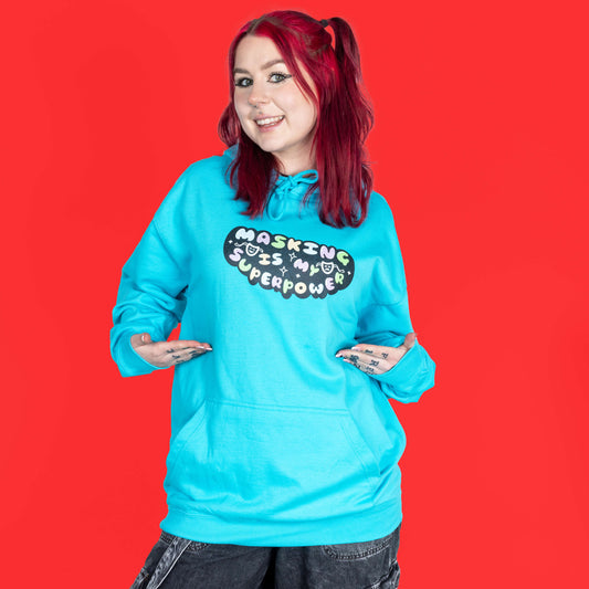 The Masking Is My Super Power Hoodie in Turquoise Surf modelled by Flo with red hair and black eyeliner on a red background. She is facing forward smiling pointing towards the hoodie graphic. The aqua blue hoodie features pastel rainbow bubble writing that reads 'masking is my superpower' with white sparkles, a happy and sad drama masks all on a black oval. Raising awareness for neurodivergent people with ADHD or Autism.