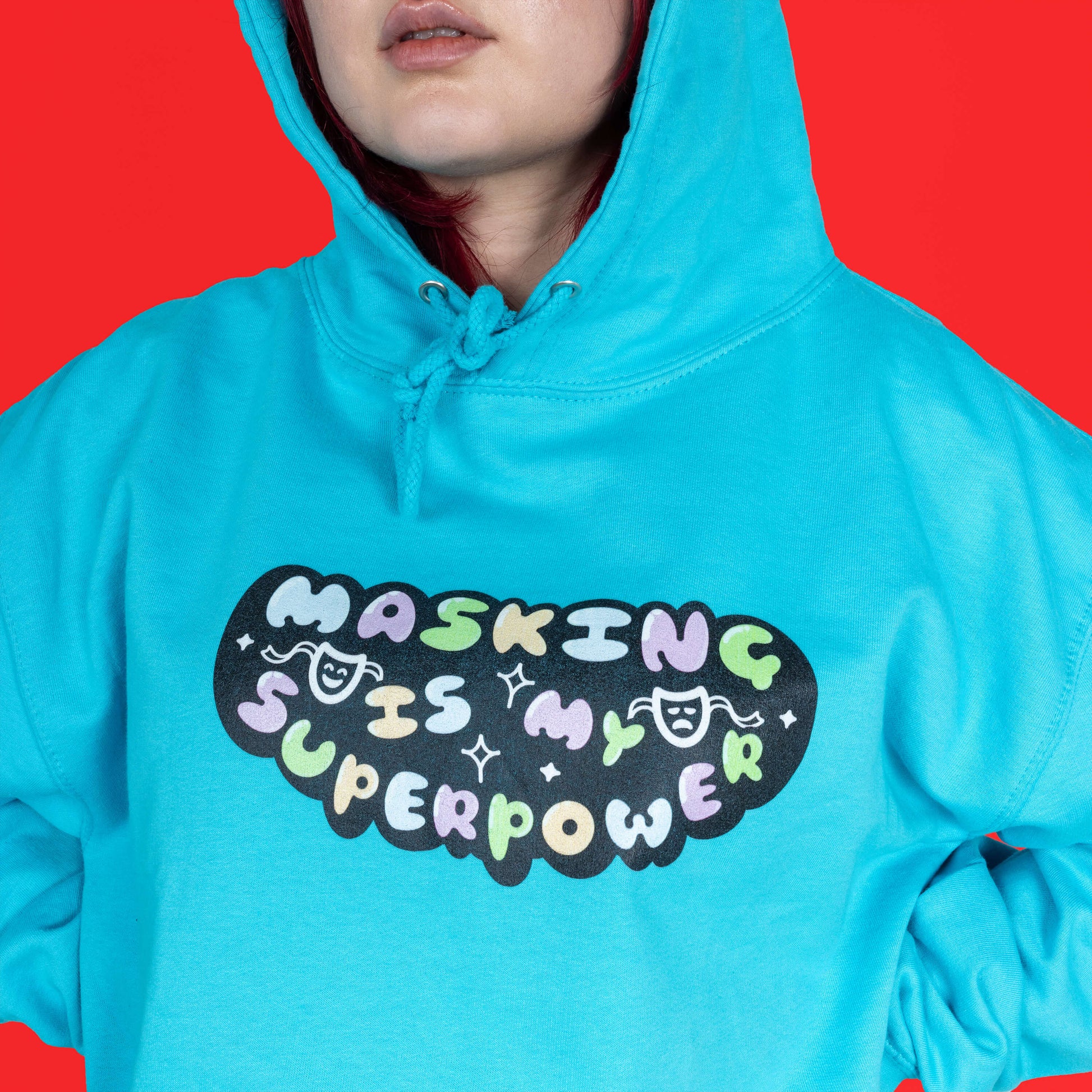The Masking Is My Super Power Hoodie in Turquoise Surf modelled by Flo with red hair on a red background. She is facing forward  with a close up of the hood up and front print. The aqua blue hoodie features pastel rainbow bubble writing that reads 'masking is my superpower' with white sparkles, a happy and sad drama masks all on a black oval. Raising awareness for neurodivergent people with ADHD or Autism.
