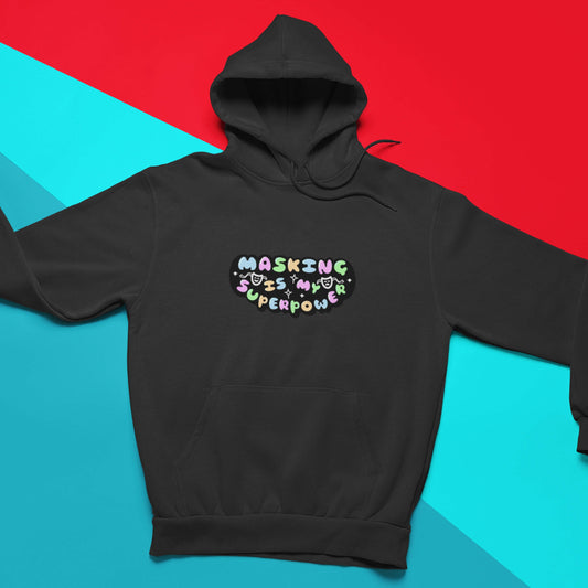The deep black Masking Is My Super Power Hoodie on a blue and red background. The black hoodie features pastel rainbow bubble writing that reads 'masking is my superpower' with white sparkles, a happy and sad drama masks all on a black oval. Raising awareness for neurodivergent people with ADHD or Autism.