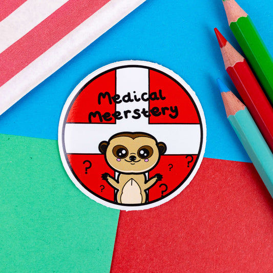A circular sticker with white and red first aid cross as the background with a cute meerkat at the bottom with big eyes and pink cheeks and has it's little arms open wide. There are black question marks around the meerkat and 'medical meerstery' written above it in black writing. It is on a red, blue and green background with coloured pencils to one side and a stripy paper bag